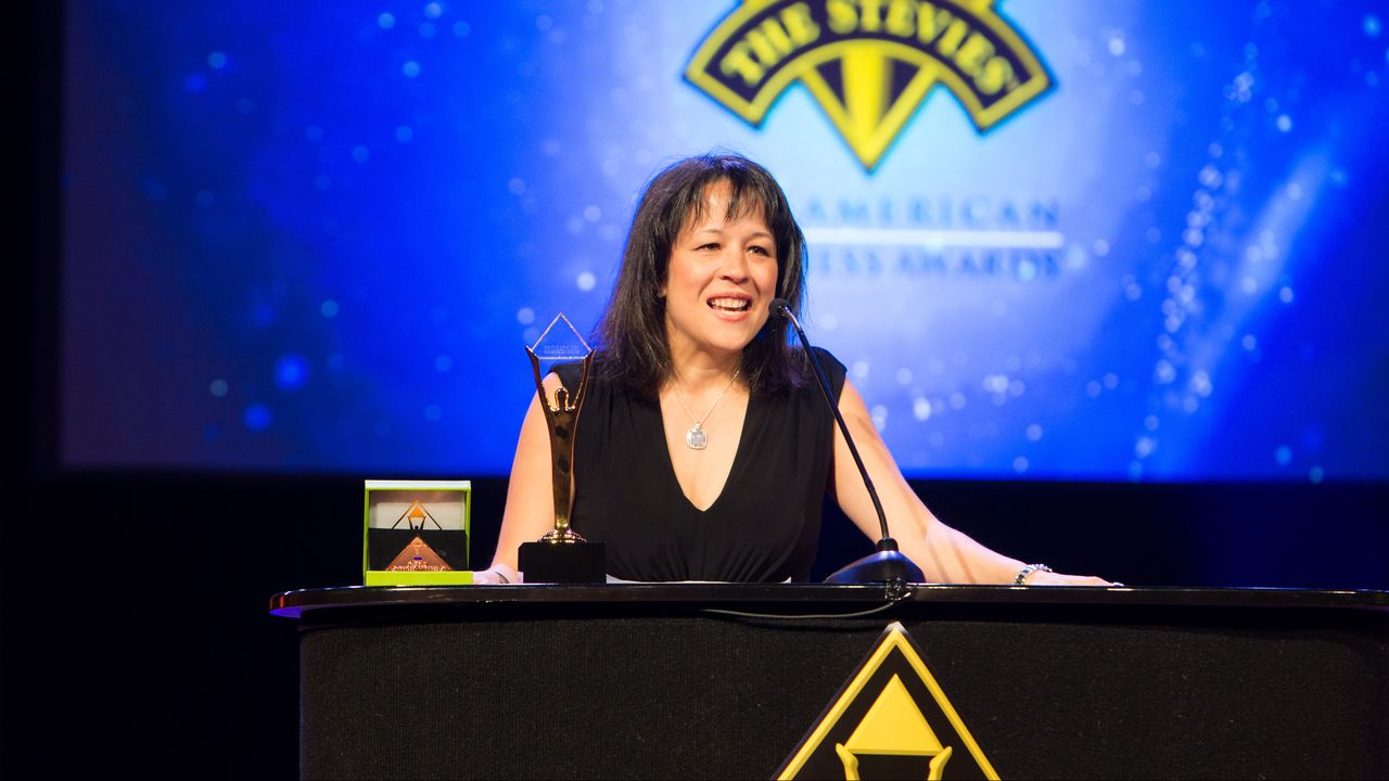 Jeannie Finks at the Stevie Awards A woman smiles while holding a trophy and speaking at the Stevie Awards ceremony.
