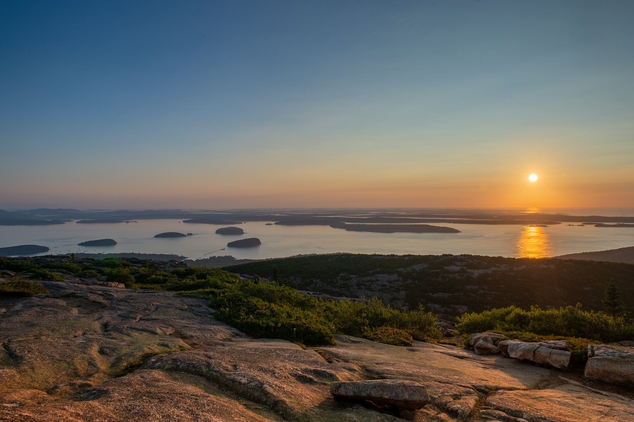 Cadillac mountain The sun rises over distant islands and calm water, viewed from a rocky hilltop with green vegetation.