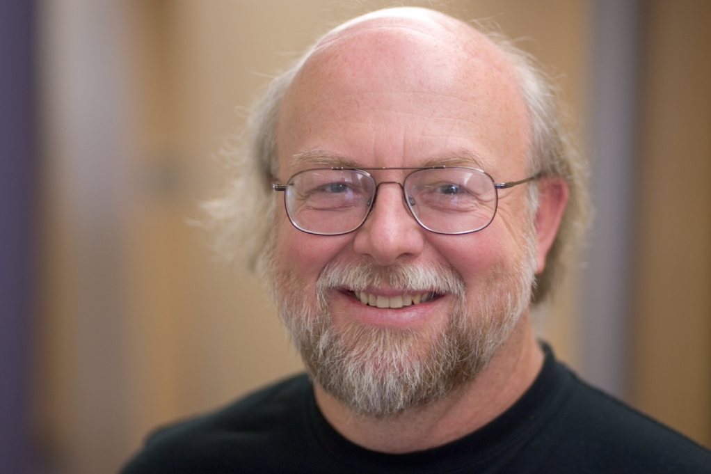 James Gosling A man with glasses and a beard smiling, wearing a black shirt, in an indoor setting.